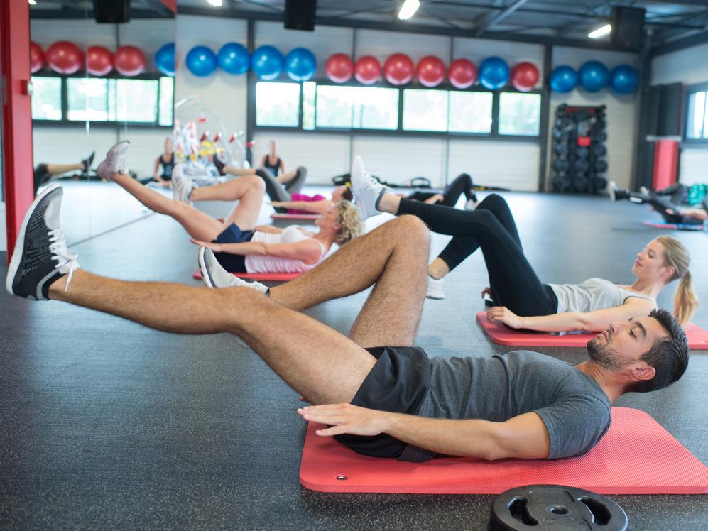 Gruppe von Personen macht Übungen auf Fitnessmatten in einem modernen Gym mit Fitnessgeräten im Hintergrund.